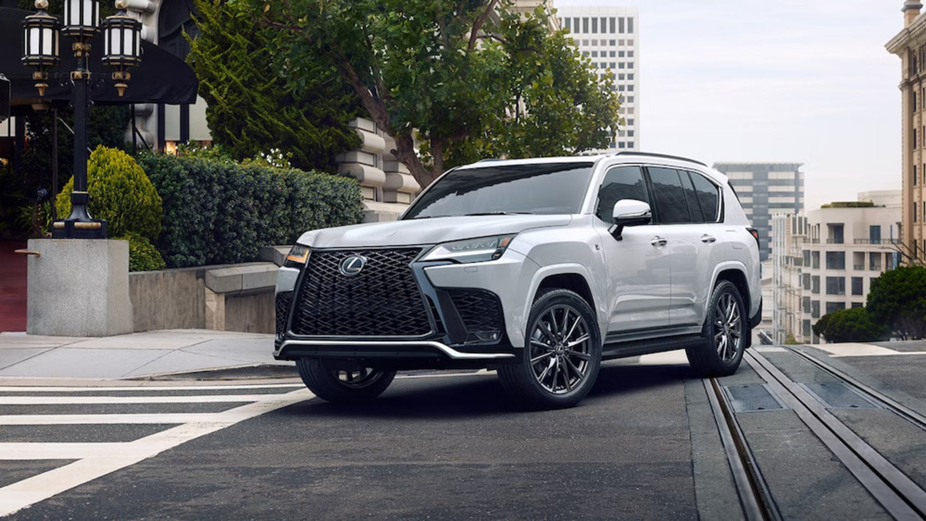 A white 2026 Lexus LX SUV turning the corner of a tree-lined city street