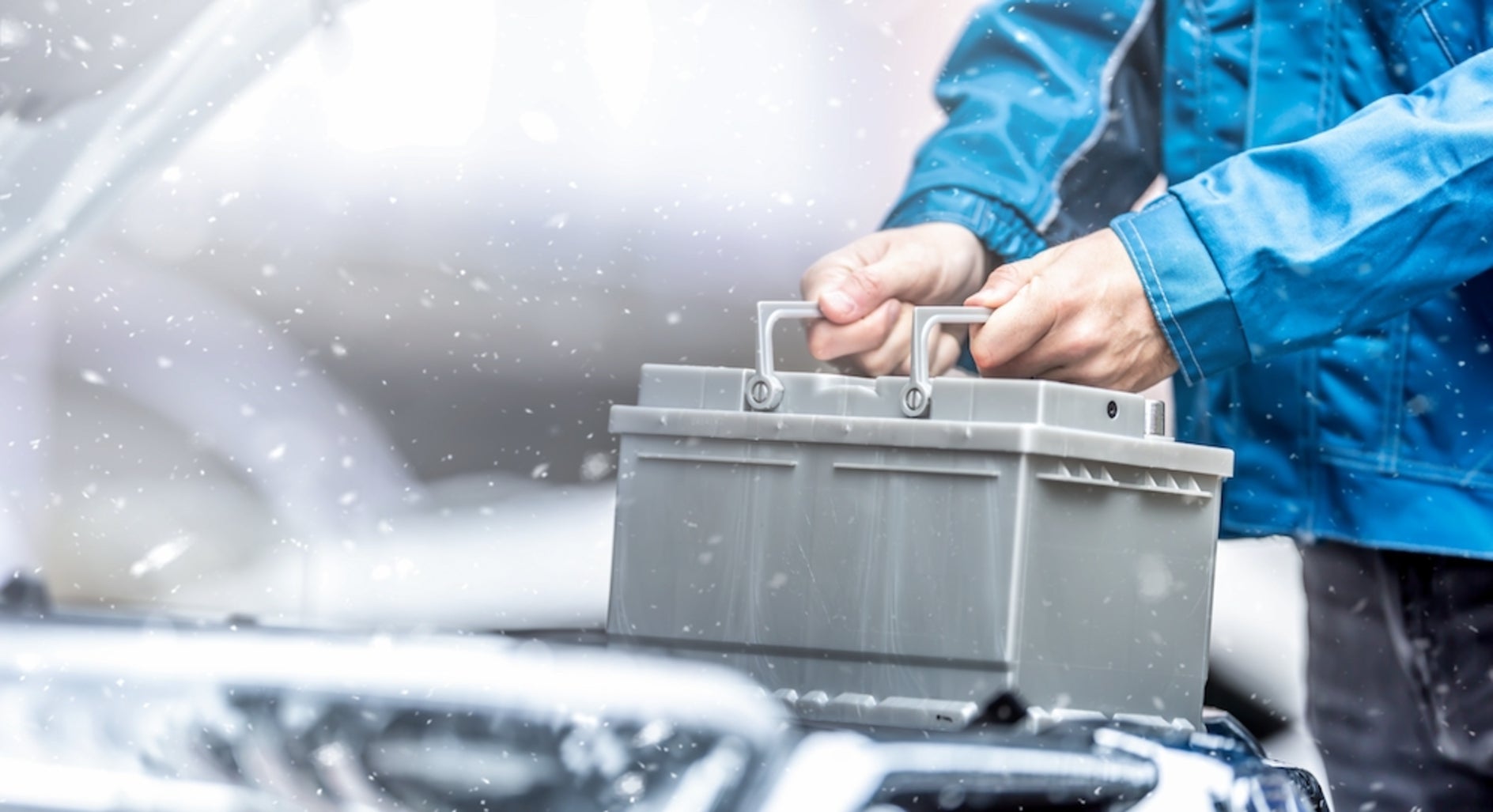 A white person in a blue jacket holding a car battery by a vehicle’s open engine, with snow falling