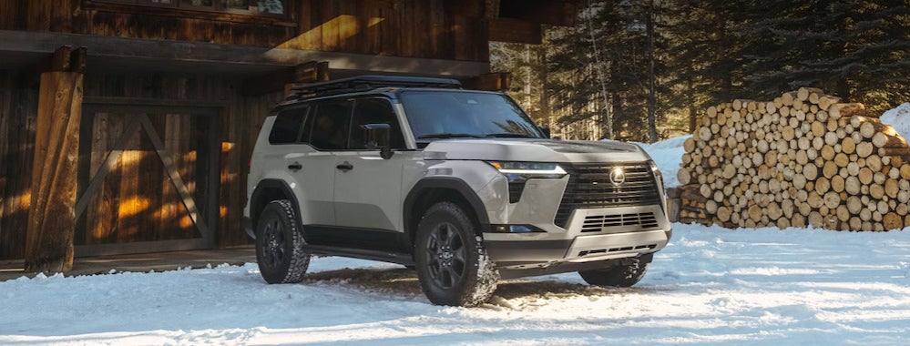 A gray 2026 Lexus GX SUV parked in front of a snowy cabin, with wood stacked in the background